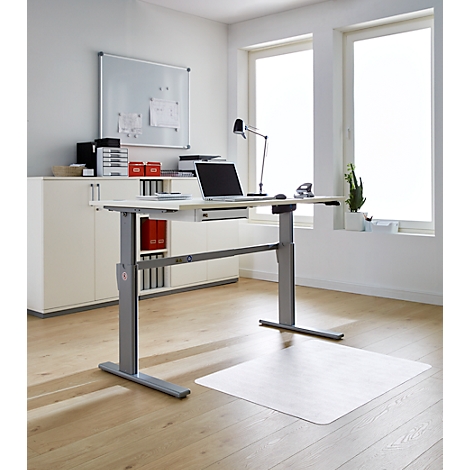 An adjustable height desk with a laptop, lamp and plants. The bright room has large windows.