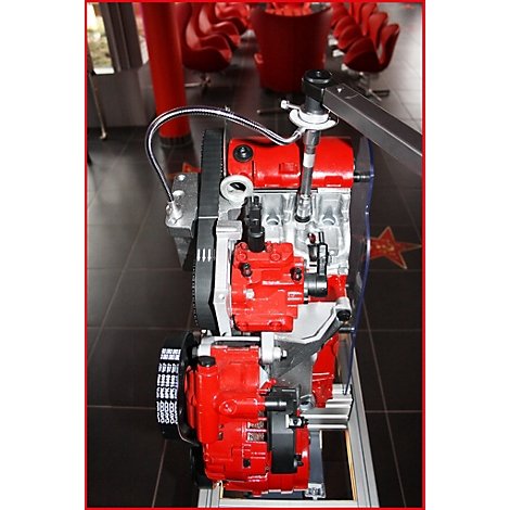 Motor im Schnitt. Rote und silberne Komponenten sind erkennbar. Der Hintergrund zeigt rote Stühle und einen Stern.