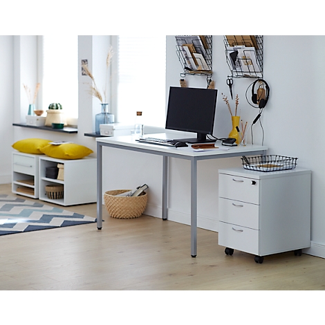 Een minimalistische werkplek met bureau, computer, opbergruimte en ladekast. Gele kussens op een kleine bank bij het raam.