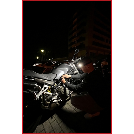 Mann repariert Motorrad im Dunkeln. Im Hintergrund ein Wohnhaus und weitere Fahrzeuge.