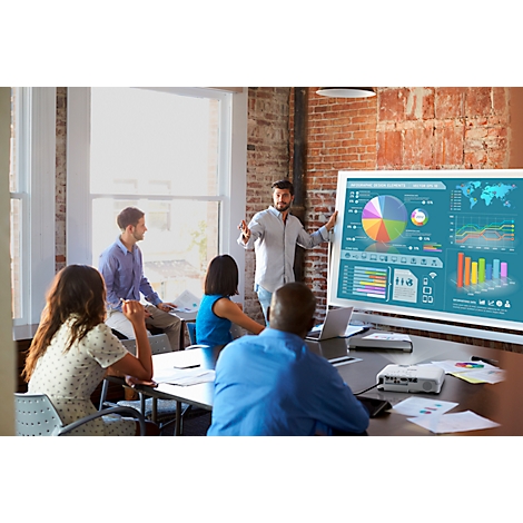 A presentation in a conference room. A man stands in front of a screen with diagrams. Other people sit at the table.