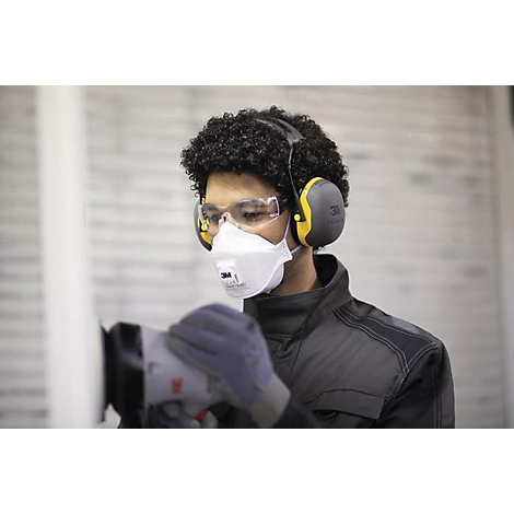 Een man met krullend haar draagt een veiligheidsbril, een masker, oorkappen en handschoenen. Hij schuurt iets met een machine.