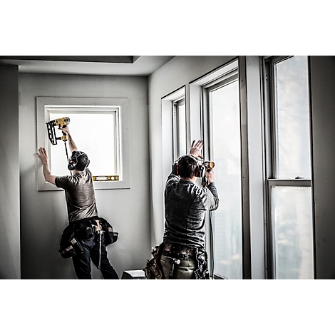 Zwei Handwerker installieren Fenster. Einer nagelt, der andere schraubt. Beide tragen Gehörschutz und Werkzeugtaschen.