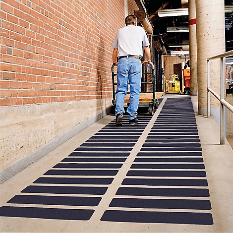 Ein Mann schiebt einen Handwagen auf einer Rampe mit schwarzen Streifen. Rote Backsteinmauer und Säule im Hintergrund.