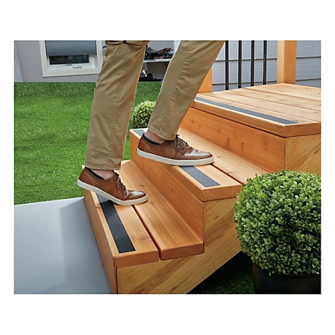 Person steigt Holztreppe hinauf. Auf jeder Stufe ein schwarzer Streifen. Auf einer Seite grüner Rasen, im Hintergrund ein Fenster.