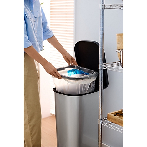 Silberfarbener Mülleimer mit schwarzem Deckel, vor einer Fensterfront und Regalen.