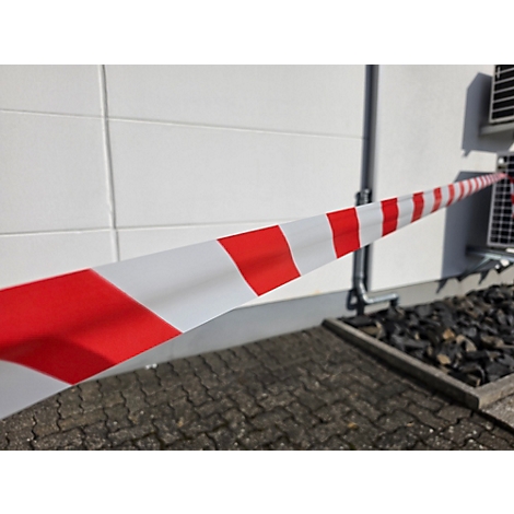 Rot-weiß gestreiftes Absperrband spannt sich diagonal vor einer weißen Hauswand.