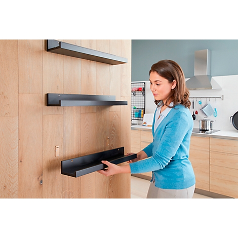 Woman installing a black wall shelf on a wooden wall. The kitchen is visible in the background.