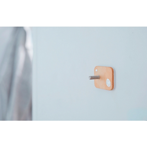 A square, wood-colored wall hook with a silver pin, attached to a white wall. To the right of it is a bright spot.