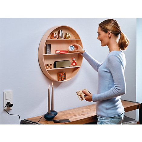 A woman places an alarm clock on a round wooden shelf. A speaker, books, and toys are on the lower shelves. Two candles are burning below.