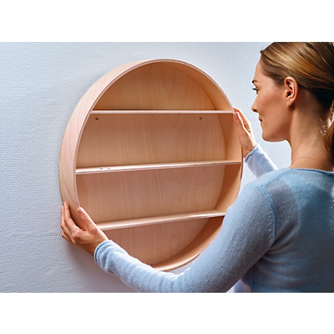 Woman holding a round wooden shelf on the wall. Shelf has three levels. Background is a whitewashed wall.