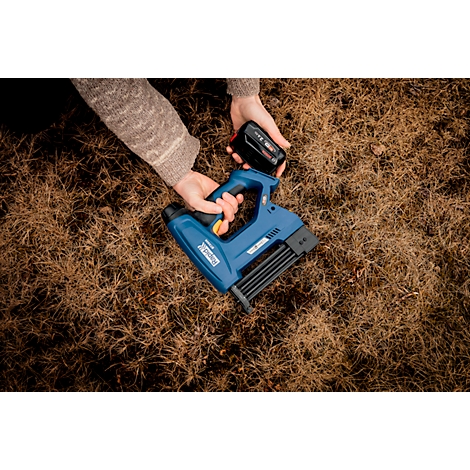 Mains tenant une agrafeuse bleue et une batterie au-dessus d'herbe sèche.