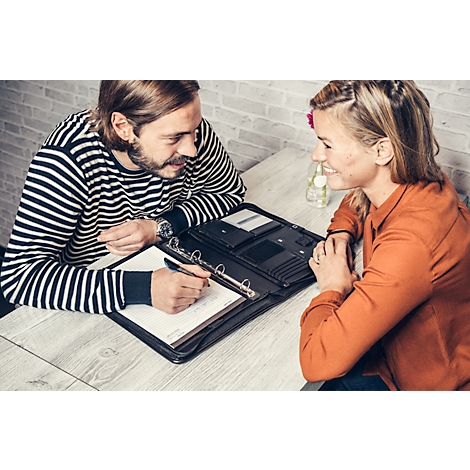 A man in a striped sweater writes in a folder while a woman sits opposite him smiling.