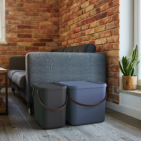 Two trash cans with lids and handles stand in front of a couch and brick wall.