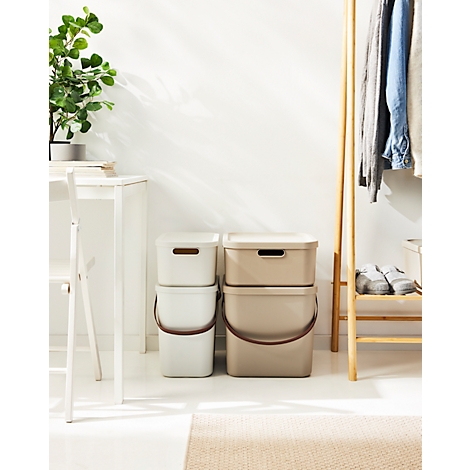 Two stacks of storage boxes with lids in white and beige, brown handles. In the background clothes rack, table and potted plant.