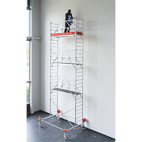 Person auf einem Gerüst arbeitet an einer weißen Wand. Graues Gerüst mit roten Akzenten und Rädern.