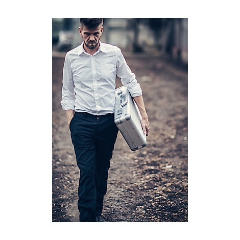 A man with a suitcase walks a path, serious look. He wears a shirt and pants.