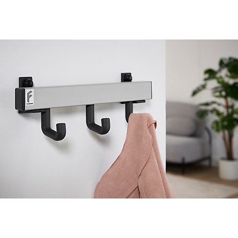 Hakenleiste mit drei schwarzen Haken, an der eine rosafarbene Jacke hängt. An der Wand hängt die Leiste.