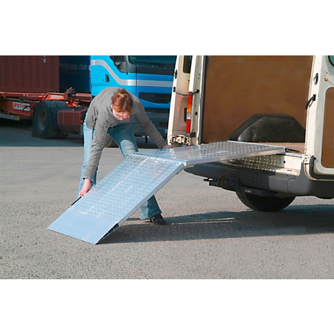 Person opening a loading ramp on a white van. A blue truck cabin can be seen in the background.