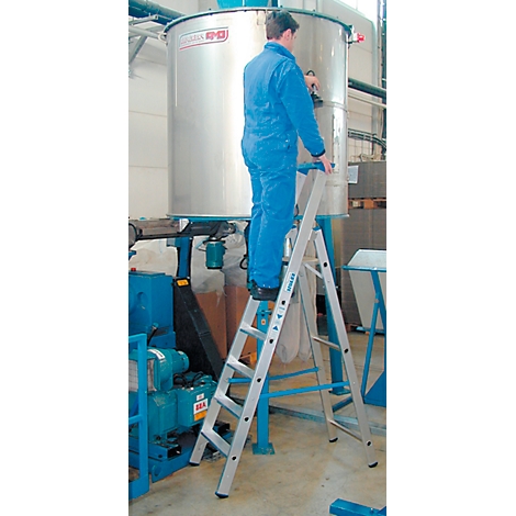 A man in blue overalls stands on a ladder in front of a large silver container.