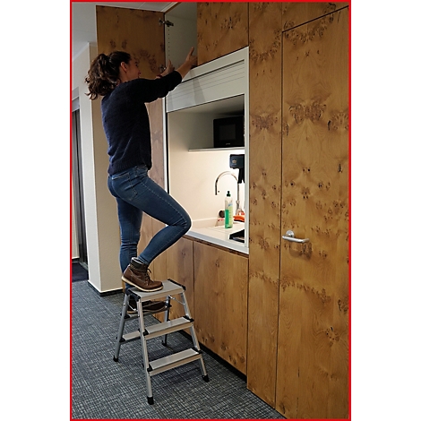 Eine Frau steht auf einer Trittleiter und öffnet einen Schrank in einer Küche.
