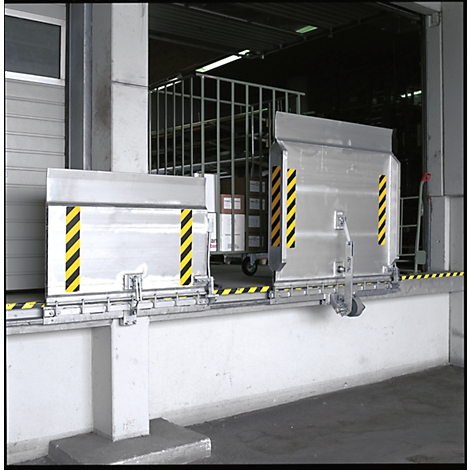 Two silver metal parts, marked yellow and black, in front of a warehouse. In the background, a wire cage cart with boxes.