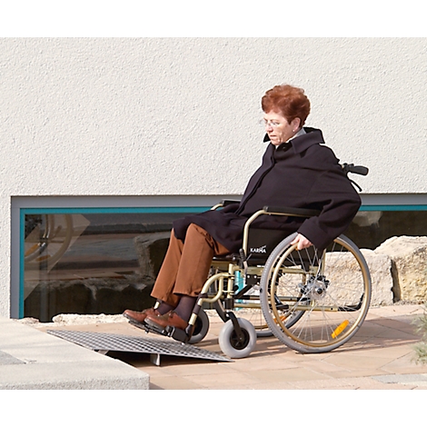 An elderly woman in a wheelchair drives over a ramp. She is wearing a black coat. In the background a window and stones.