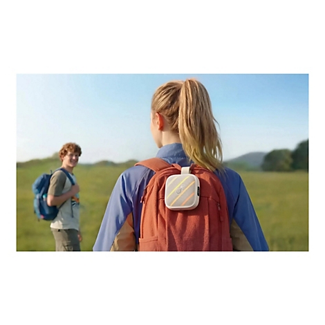 Eine Wanderin mit Rucksack und tragbarem Lautsprecher auf einer sonnigen Wiese; ein Wanderfreund steht im Hintergrund.