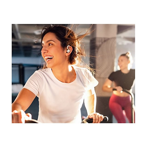 Eine lächelnde Frau trainiert auf einem Heimtrainer im Fitnessstudio und trägt weiße In-Ear-Kopfhörer.