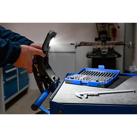 Een monteur die een zwarte werklamp met helder licht in de werkplaats vasthoudt. Op de achtergrond gereedschap, blauwe doos met opzetstukken.
