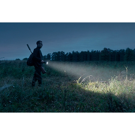 Person with rifle and backpack in field at night, flashlight illuminating the way.