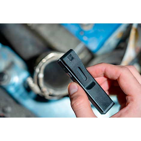 A hand holds a black, rectangular tool. In the background, a pipe with a metal ring.