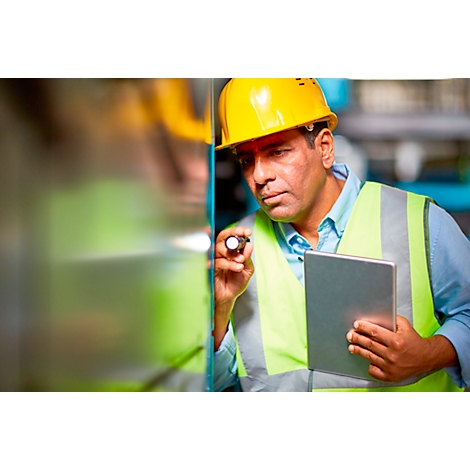 A man with helmet and vest inspects a machine with a tablet and flashlight.