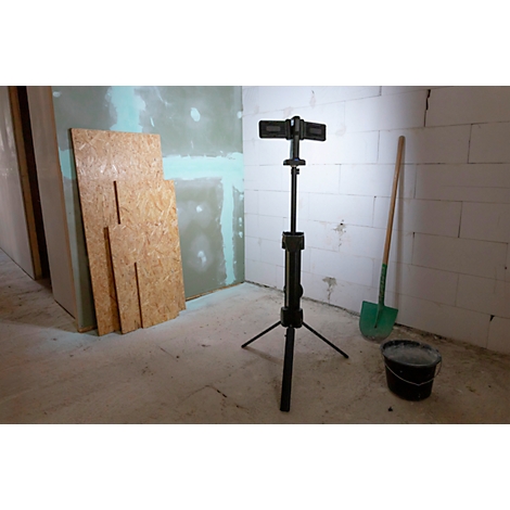 In einem Rohbau steht ein Baustrahler auf einem Stativ, daneben eine Schaufel, ein Eimer und Holzplatten.