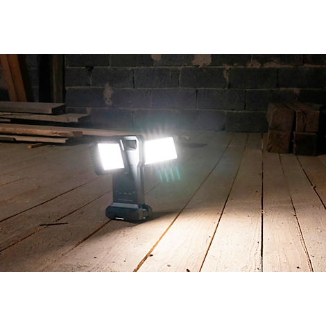 Eine schwarze, batteriebetriebene Baulampe leuchtet hell auf Holzboden. Im Hintergrund eine Steinwand.