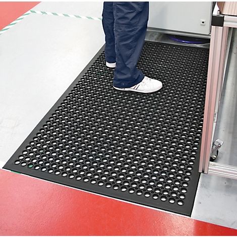 Person standing on a black perforated mat with round holes. The floor is red and gray. Feet in white shoes.