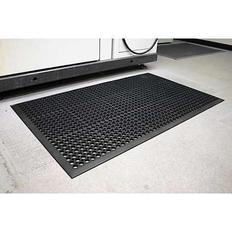 Black rubber mat with circular holes lies in front of a machine on a concrete floor.