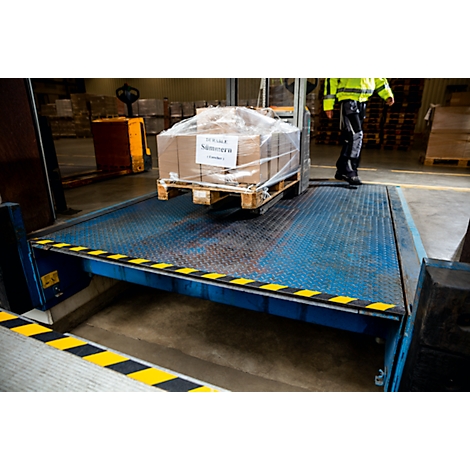 A worker in work clothes stands next to a loading ramp on which a package stands on a pallet.