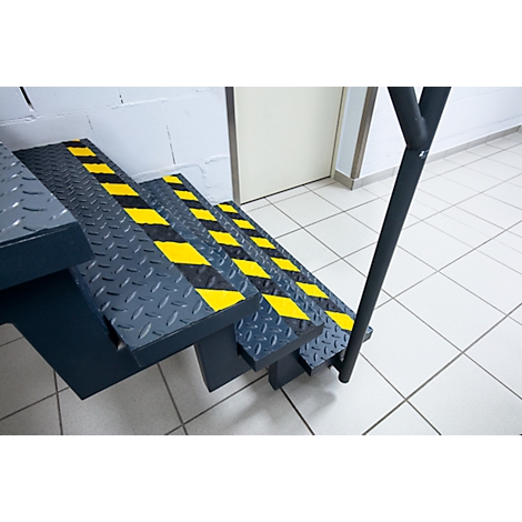 Steel staircase with yellow and black warning stripes. Handrail on the right, door and tiles in the background.