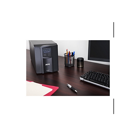 Un onduleur APC noir est posé sur un bureau marron. À côté, un moniteur, un porte-stylo, un clavier et un stylo.