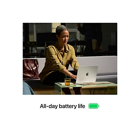 Frau sitzt an Tisch mit Laptop, im Hintergrund weitere Person. Auf dem Bildschirm ist das Apple-Logo zu sehen. Auf dem Tisch steht ein Glas.