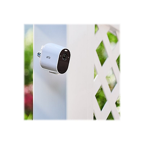 Weiße Arlo-Überwachungskamera, an einer Wand befestigt, mit einem dahinterliegenden Gitterzaun.
