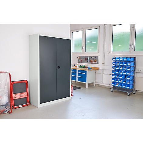 Armario metálico gris con puertas dobles en taller junto a un taburete plegable rojo. Banco de trabajo y estanterías en el fondo.