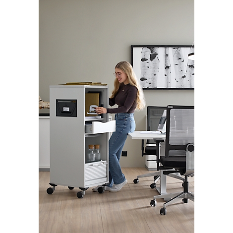 Jeune femme ouvrant un meuble roulant gris dans un bureau, sourit et en sort quelque chose.