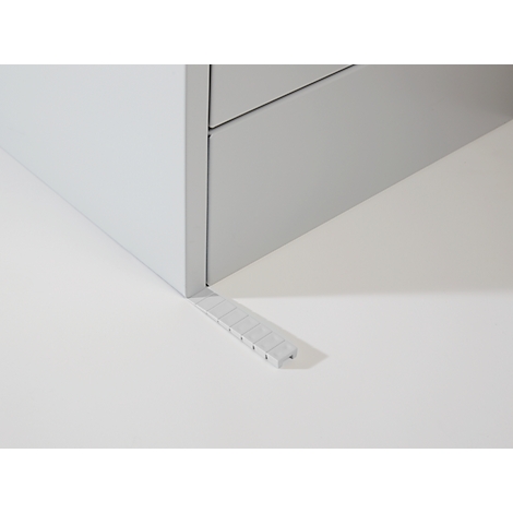 Un pied de meuble gris avec une cale. La cale est positionnée en biais sous le pied du meuble.
