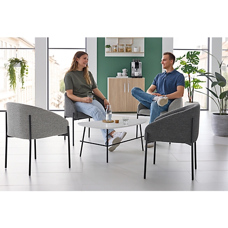Two people sit in a waiting area furnished with gray armchairs and a white table. In the background is a coffee bar.