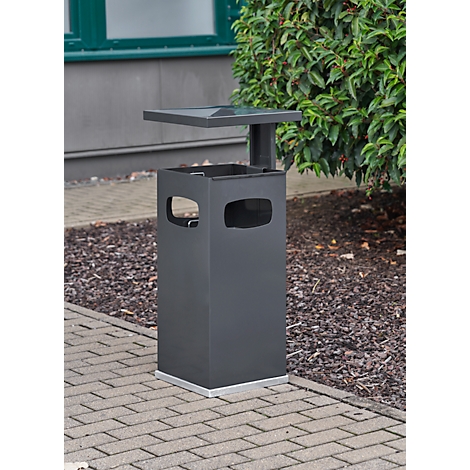 Grey outdoor trash bin with shelf, rectangular with cutouts. Standing on paving stones next to green areas.