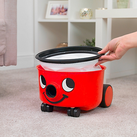 Aspirateur rouge Henry avec filtre. Une main insère le filtre. Sur tapis gris, étagère en arrière-plan.