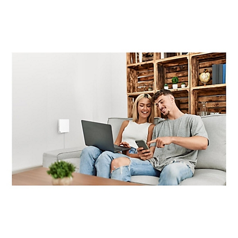 Ein lächelndes Paar sitzt auf einem Sofa, schaut auf ein Smartphone und einen Laptop. An der Wand ist ein weißes Gerät.