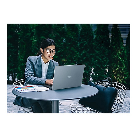 Junge Frau arbeitet mit Laptop an Tisch im Freien, lächelnd. Im Hintergrund Grünpflanzen und eine Aktentasche.